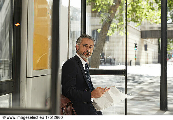 Confident businessman with newspaper waiting at bus stop