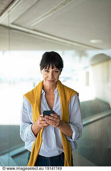 Confident businesswoman holding smart phone in front of glass wall