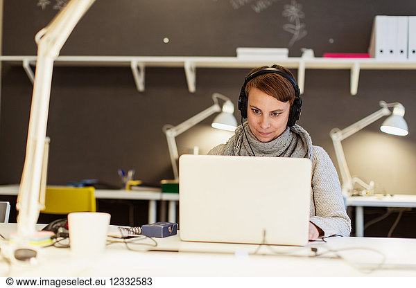 Confident businesswoman listening to headphones while using laptop at creative office
