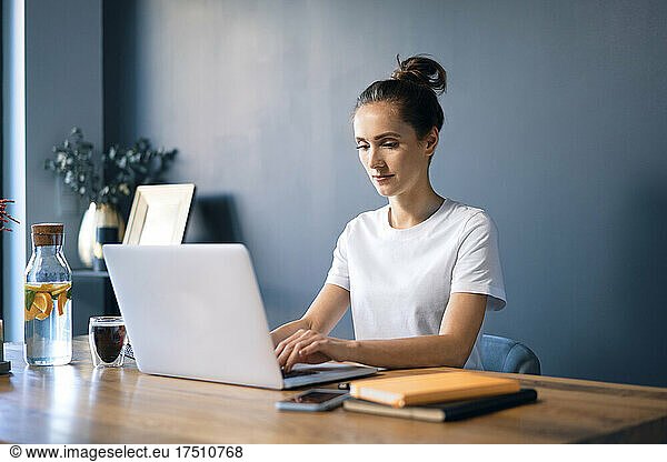 Confident businesswoman using laptop on desk against wall in home office
