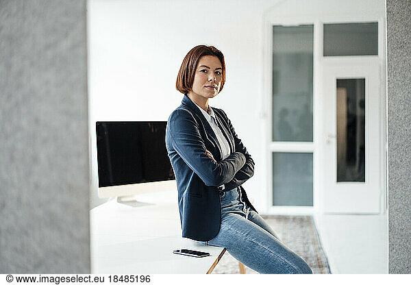 Confident businesswoman with arms crossed sitting on desk