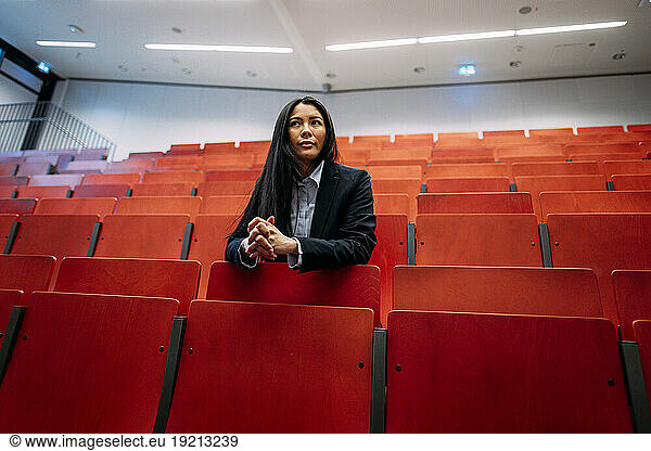 Confident businesswoman with hands clasped leaning on red seats in auditorium
