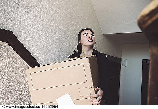 Confident delivery woman with cardboard box looking up against wall