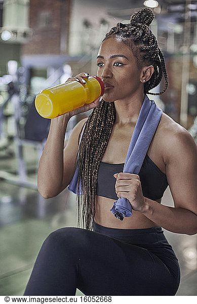 Confident female athlete with braided hair drinking water while standing in gym