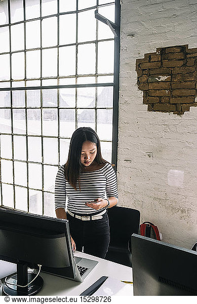 Confident female computer programmer using technologies standing by desk at creative office