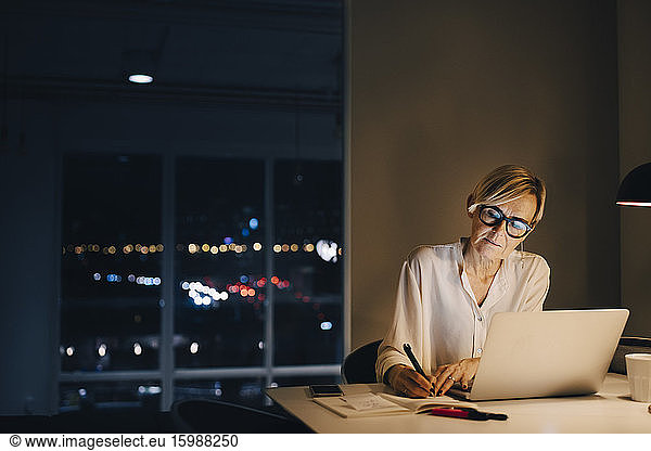 Confident female entrepreneur writing in book while sitting with laptop at office desk