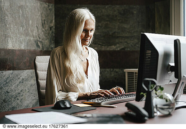 Confident female financial advisor with long white hair using computer at desk in office