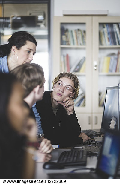 Confident female mature teacher discussing with male students while looking at computer monitor in classroom