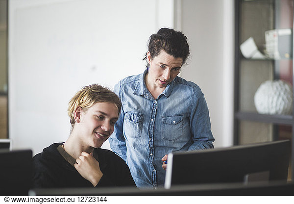 Confident female mature teacher looking at computer monitor while standing by young student in classroom