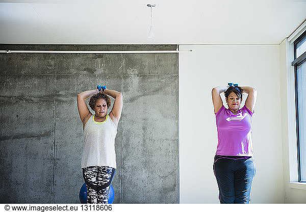 Confident friends using dumbbells while exercising against wall in yoga studio
