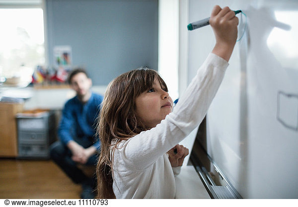 Confident girl drawing on blackboard while teacher sitting in background
