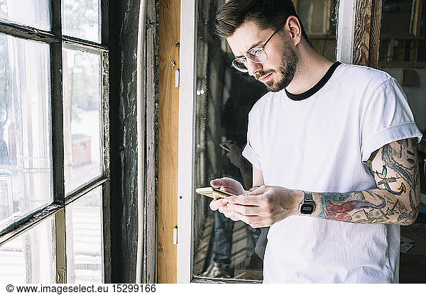 Confident male hipster hacker text messaging through smart phone by window at creative office
