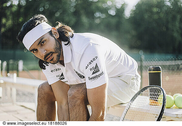 Confident man wearing headband while sitting on bench at tennis court