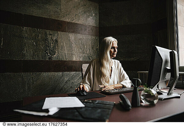 Confident mature businesswoman with long white hair using computer at desk in office