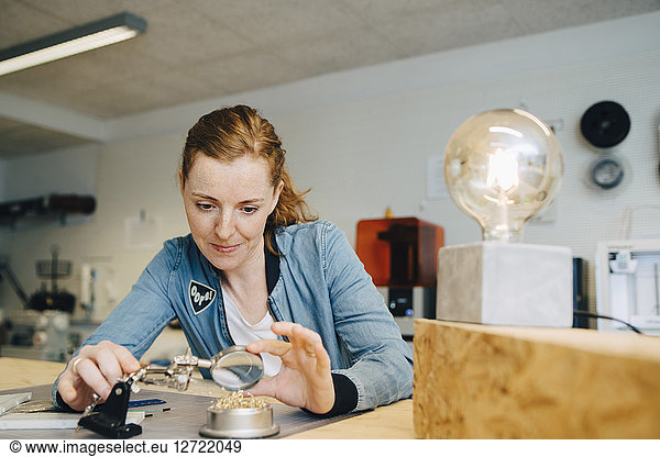 Confident redhead female engineer looking through magnifying glass at workbench in creative office