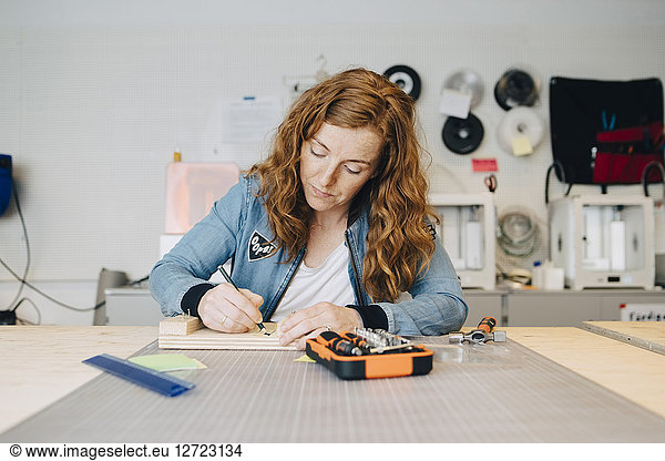 Confident redhead female engineer writing on wood at workbench in creative office