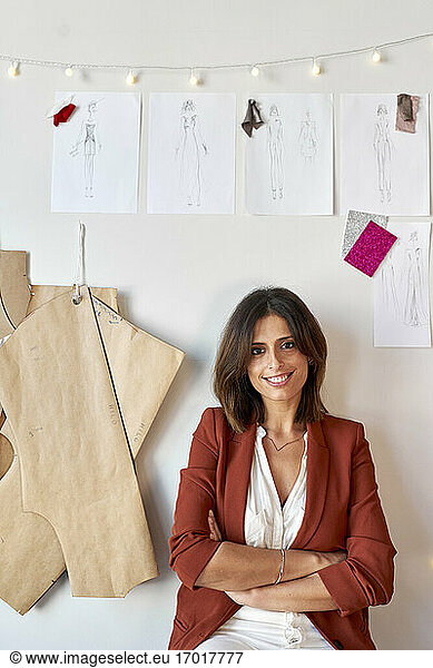 Confident smiling fashion designer sitting with arms crossed below her designs and patterns on white wall at atelier