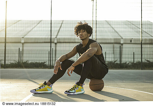Confident sportsman sitting on basketball at sports court