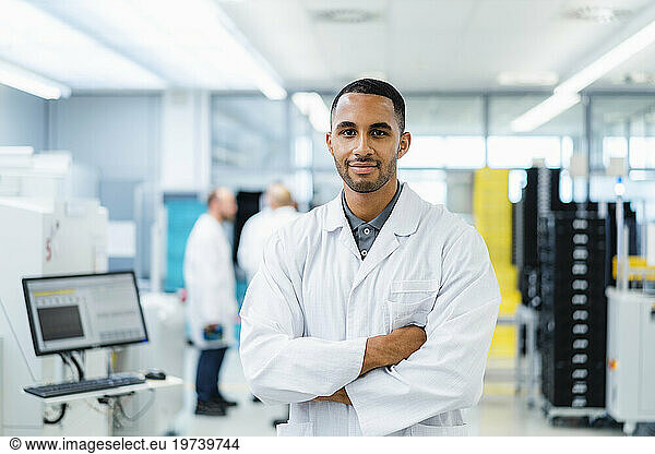 Confident technician smiling with arms crossed while colleagues are talking in electronics factory