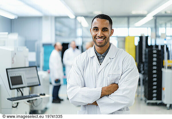 Confident technician smiling with arms crossed while colleagues are talking in electronics factory