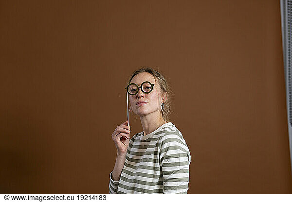 Confident woman holding eyeglasses prop over brown background