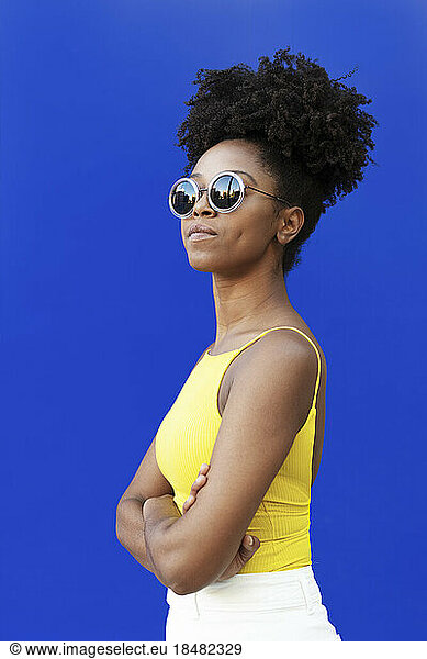 Confident woman wearing sunglasses with arms crossed by blue background