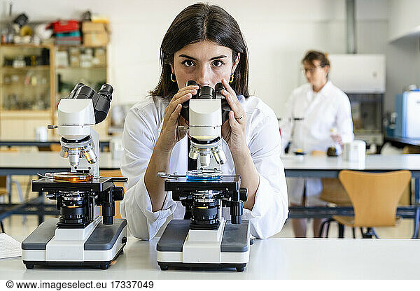 Confident young female scientist leaning on desk with microscope in laboratory