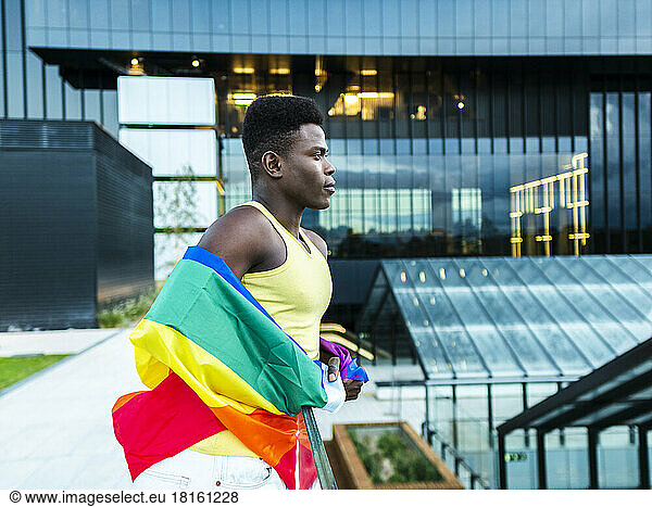 Confident young man wrapped in rainbow flag leaning on railing by modern glass buildings