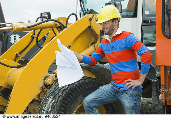 Construction worker holding construction plan in front of excavator