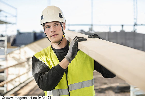 Construction worker looking away while carrying wooden plank at site