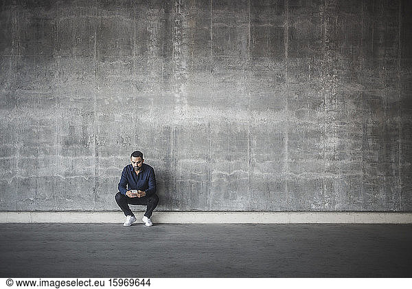 Contemplating man using cell phone while crouching against gray wall