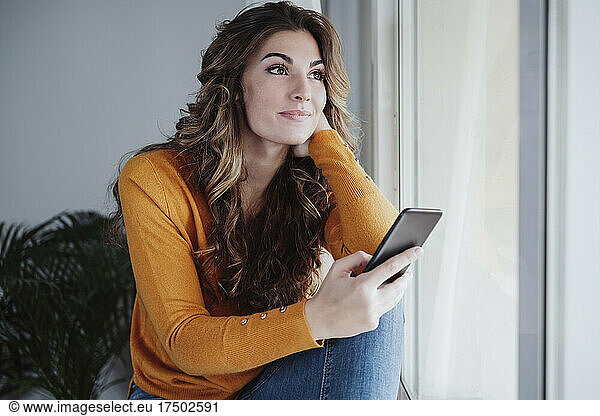 Contemplating woman with smart phone sitting near window at home