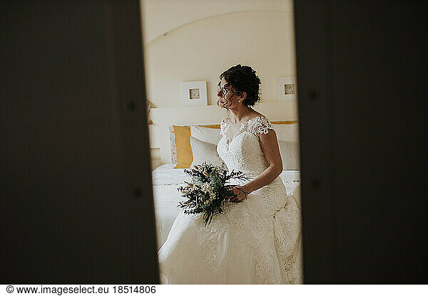 Contemplative bride sitting with bouquet of flowers on bed