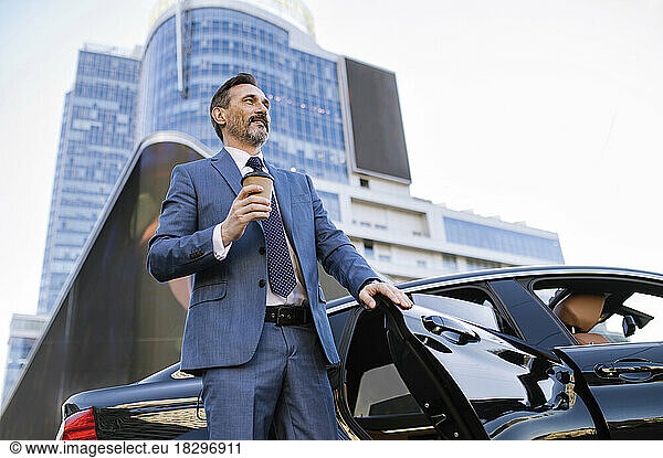Contemplative businessman with disposable coffee cup standing by car outside building