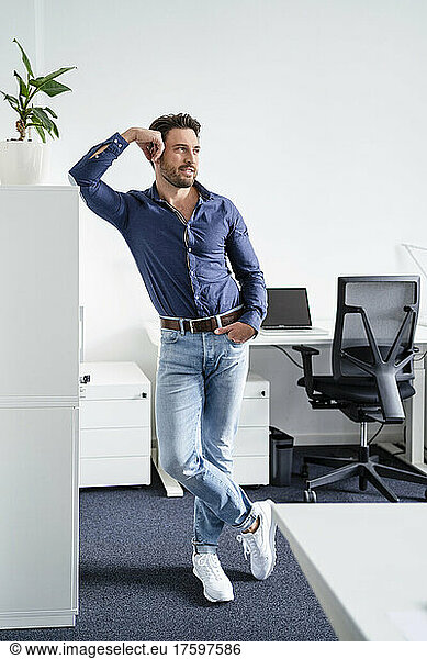 Contemplative businessman with hand in pocket leaning on cabinet at office