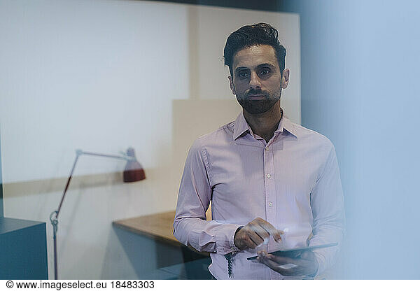 Contemplative businessman with tablet PC standing in office seen through glass
