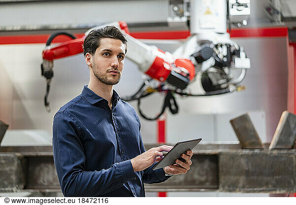 Contemplative engineer holding tablet PC in front of robotic arm
