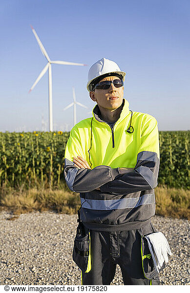 Contemplative engineer standing with arms crossed in front of wind turbines