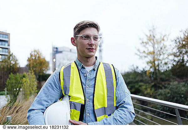 Contemplative engineer wearing eyeglasses holding hardhat under arm
