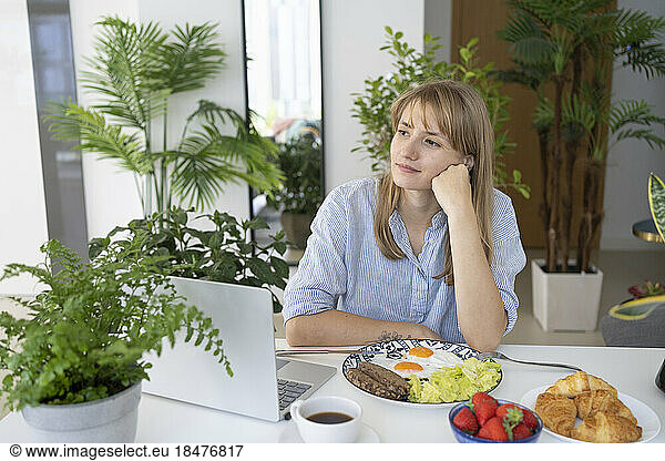 Contemplative freelancer sitting with breakfast and laptop at desk