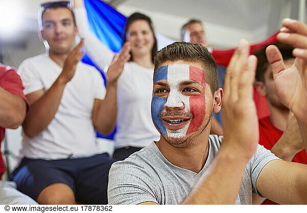 Contemplative man with French Flag painted on face clapping with fans in stadium