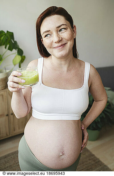 Contemplative pregnant woman holding glass of green smoothie at home