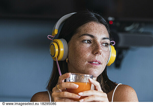 Contemplative woman wearing headphones holding glass of juice at home