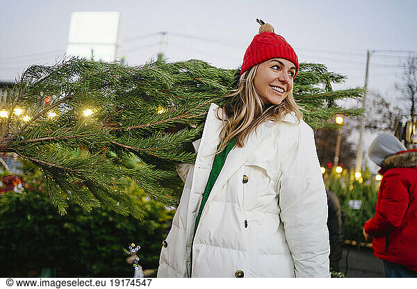 Contemplative woman wearing knit hat carrying Christmas tree at market