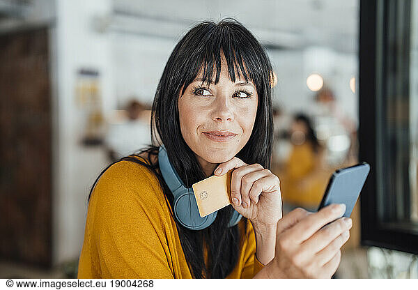 Contemplative woman with with headphones around neck holding credit card and smart phone
