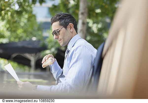Corporate businessman eating lunch and reading paperwork on park bench