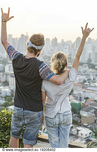 Couple gesturing horn sign in front of cityscape at sunset