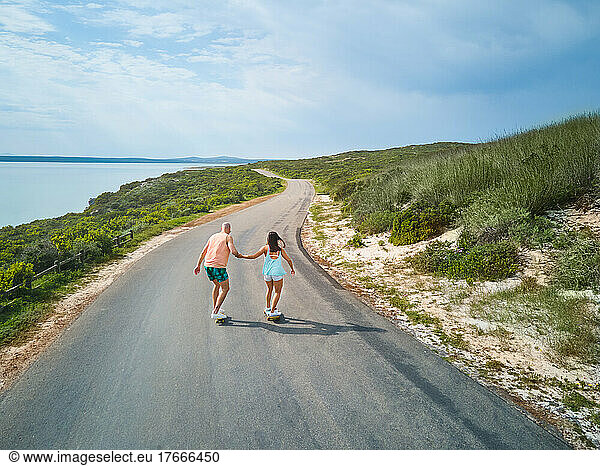 Couple holding hands and skateboarding on sunny ocean road