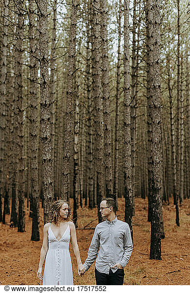 couple holding hands looks at each other amid pine trees in forest.