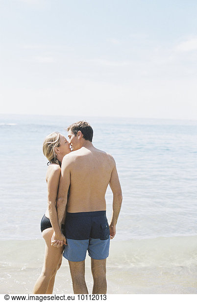 Couple in swimwear standing on a sandy beach by the ocean  kissing.